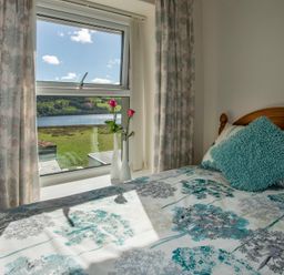 A bedroom with a wooden bed a blue cushion floral curtains and two vases with pink flowers by the window at 17 Victoria Terrace - Perthi in Nantlle