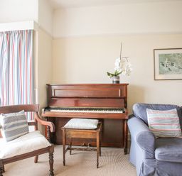 A living room with a wooden piano a wooden chair a wooden piano stool a blue couch with cushions and striped curtains at Windygates in Rhosneigr