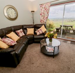 A living room with a black leather corner sofa and cushions a glass coffee table and sliding glass doors opening to a patio at Forge Cottage in Rothbury