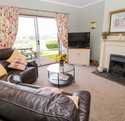 A living room with leather sofas a round glass coffee table a television on a stand a fireplace and sliding glass doors with floral curtains at Forge Cottage in Rothbury