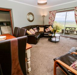 A living room with a brown leather sectional sofa and a wooden dining table with chairs next to large sliding glass doors with floral curtains at Forge Cottage in Rothbury