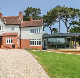 A house with a gravel driveway and a lawn with trees behind at The Coach House in Teignmouth