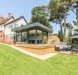 A modern house with a glass extension and wooden deck in a garden with patio furniture and a barbecue at The Coach House in Teignmouth