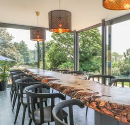A dining room with a long wooden table surrounded by black chairs and large windows showing greenery at The Coach House in Teignmouth