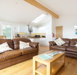 A living room with two brown leather sofas and a wooden coffee table in front of a kitchen with white cabinets at 3 Valley View in Lanreath
