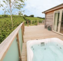 A wooden deck with a hot tub a garden hose and glass doors opening to a wooden house with green hills in the background at 3 Valley View in Lanreath