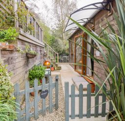 An outdoor walkway with a wooden fence and gate leading to a wooden building with an open door and plants along the sides at Wellinghill Lodge in Cheltenham