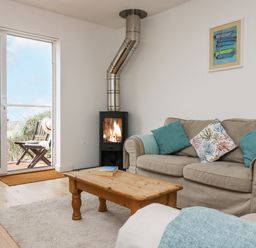A living room with a beige sofa with blue and patterned cushions a wooden coffee table a fireplace and a sliding glass door leading to an outdoor seating area at Sunset in St Just