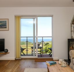 A living room with a sliding glass door opening to a balcony overlooking green hills and the sea at Sunset in St Just