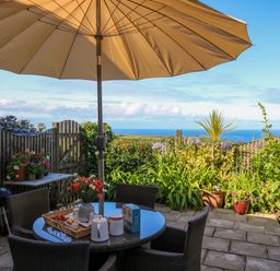 A patio with a round table and chairs under a large umbrella with plants and a view of the sea at Sunset in St Just