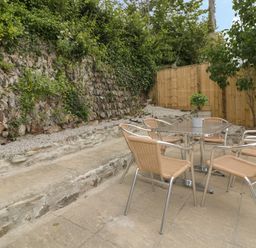 An outdoor patio with a metal table and four wicker chairs next to a stone wall and wooden fence at Avalon in St Agnes