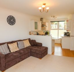An open plan living room with a brown sofa with cushions a wall clock and a kitchen area at Seashore in Woolacombe