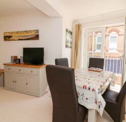 A dining area with a table covered by a tablecloth and four chairs next to a cabinet with a television at Seashore in Woolacombe