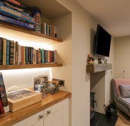 A room with wooden shelves holding books and board games a TV mounted above a fireplace and a gray chair with a pink blanket at St Marys Cottage in Windermere