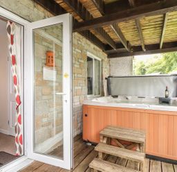 An outdoor covered patio with a wooden hot tub and steps next to a glass door leading to a room with a red bedspread at Valley Lodge 52 Callington