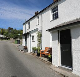 A white exterior of a cottage with a black door and wooden bench on a narrow road at Bryn Teg Cottage in Menai Bridge