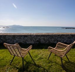 Two outdoor wicker chairs on grass facing a stone wall with a view of the sea and distant hills at Stabl Bach in Llanfaethlu