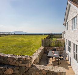 A patio with a table and chairs beside a white house overlooking a fenced grassy area and the sea at Stabl Bach in Llanfaethlu