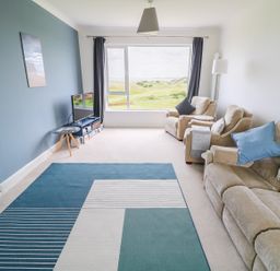 A living room with beige sofa and chairs a blue and white rug a television near a large window with a view of green hills at Seacoast Cottage in Castlerock