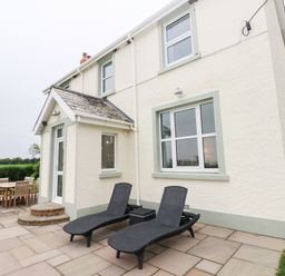 An outdoor patio area with two black lounge chairs a small table and a wooden dining table with chairs outside a two-story house at Copybush Cottage in Lawrenny