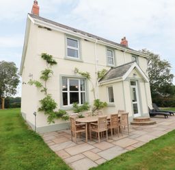 The exterior of a house with outdoor dining table and chairs on a patio and a lawn with trees at Copybush Cottage in Lawrenny