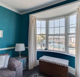 A living room corner with teal walls a gray sofa a side table with a lamp a window with white curtains and a cushioned wooden bench at Sailor's Watch in Weymouth