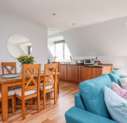 A dining area with wooden chairs and a table with flowers next to a kitchen with wooden cabinets and a blue sofa at Isallt Cove in Trearddur Bay