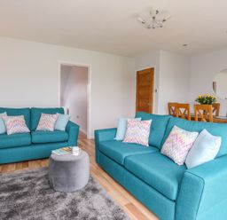A living room with two blue sofas and a round gray ottoman next to a dining table with wooden chairs at Isallt Cove in Trearddur Bay