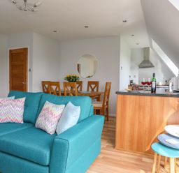 A living area with a turquoise sofa and multicolored pillows a wooden dining table with chairs and a round mirror on the wall a kitchen area with a wooden counter a microwave and wine glasses at Isallt Cove in Trearddur Bay