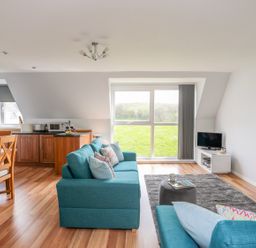 A living room with blue sofas a gray rug a small gray ottoman and a television on a white stand at Isallt Cove in Trearddur Bay