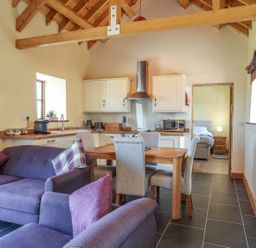 An open-plan kitchen and living room with purple sofas a wooden dining table with grey chairs kitchen cabinets and a doorway leading to a bedroom at Y Bwthyn in Brynsiencyn