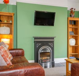 A living room with a green wall a mounted television a fireplace brown leather sofas and wooden shelves at Sefton House in Keswick