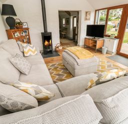 A living room with a beige sectional sofa a wood burning stove television on a stand and wooden framed glass doors at The Old Farm Shop in Narberth