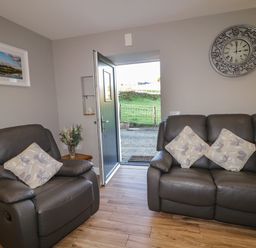 A living room with two grey leather sofas with floral cushions an open door showing outside a grassy area a clock on the wall and a framed picture at Lough Island Reavy Cottage in Kilcoo Mourne Mountains Newry County Down