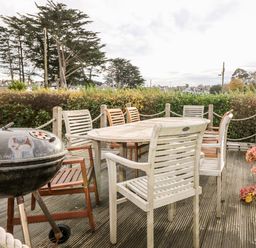 An outdoor patio with a wooden table surrounded by chairs and a charcoal barbecue grill on a deck with plants and trees at Castell in Abersoch