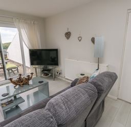 A living room with a grey sofa a glass coffee table a television and sliding glass doors leading to an outdoor patio at Sunset Sands in Deganwy