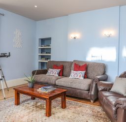 A living room with blue walls a brown sofa and armchair wooden coffee table telescope and patterned curtains at Gadlys House - Waters Edge Beaumaris