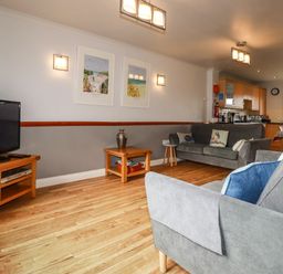 A living room with gray sofas a wooden coffee table a television on a wooden stand and a kitchen in the background at Sunflower Cottage in St Issey