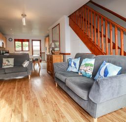 A living room with two grey sofas with blue and patterned cushions a wooden staircase and a dining area in the background at Sunflower Cottage in St Issey