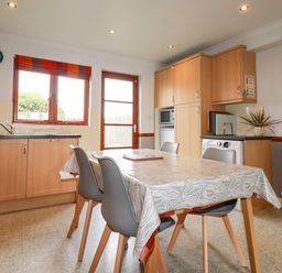 A kitchen with wooden cabinets a dining table with chairs and a door leading outside at Sunflower Cottage in St Issey