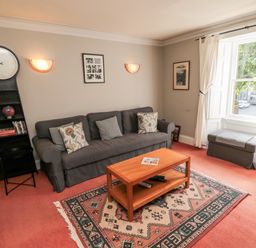 A living room with a grey sofa, wooden coffee table, patterned rug, tall black clock shelf, and a window with white curtains at Simonside Apartment in Rothbury