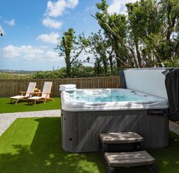 An outdoor area with a hot tub, two lounge chairs, and a fence with trees behind at Bear Cottage in Chacewater near Truro