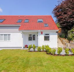 A house with a red tiled roof white walls windows and a green lawn with plants and a bench outside at The Crest in Helston