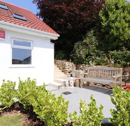 A garden area with plants a wooden bench stone steps and a white house with a red tiled roof at The Crest in Helston