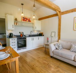 A kitchen with white cabinets wooden dining table set with bowls and glasses beige sofa with cushions wooden beams on ceiling at The Lodge at Mortons in Hankerton