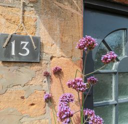 A house number 13 sign on a stone wall next to a window with textured glass and purple flowers in front at Abbey Cottage Montacute