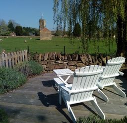 Two white wooden chairs and a small table on a wooden deck overlooking a field with sheep and a stone building at Abbey Cottage Montacute