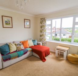 A living room with a sofa with cushions and throws a side table and a pouf by a window with floral curtains at Sea-u-Soon in Runswick Bay