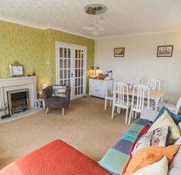 A living room with a sofa covered in colorful cushions and a blanket a dining table with six chairs a small armchair next to a fireplace and a cabinet at Sea-u-Soon in Runswick Bay