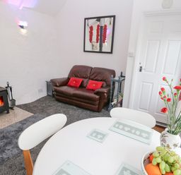 A room with a round white table with chairs a fruit bowl and flower vase a brown leather sofa and a wood burning stove at Toadstools in Rame Cross near Penryn
